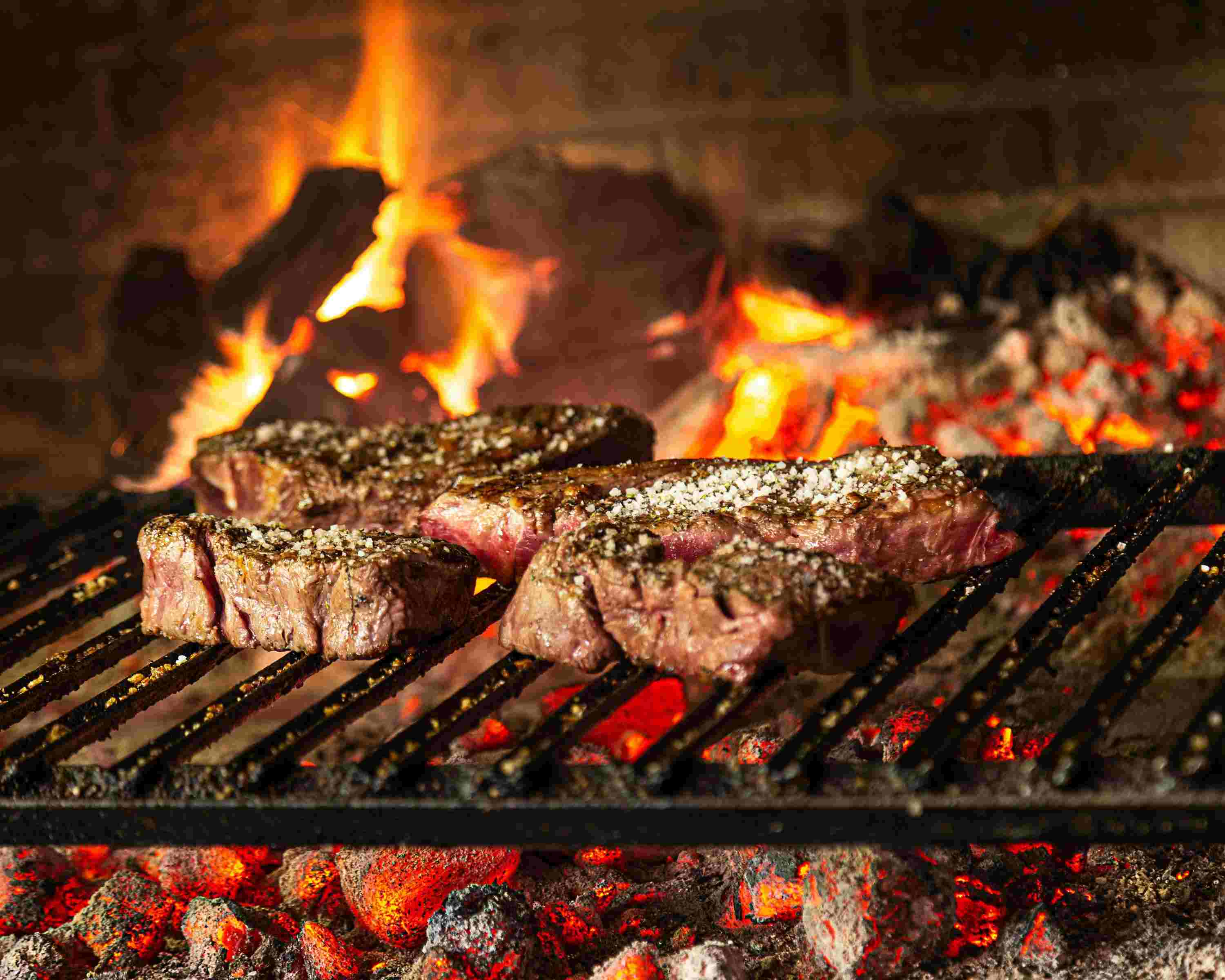 Close-up of glowing hardwood charcoal beneath a grill grate with a beautifully seared steak above the coals and realistic smoke rising