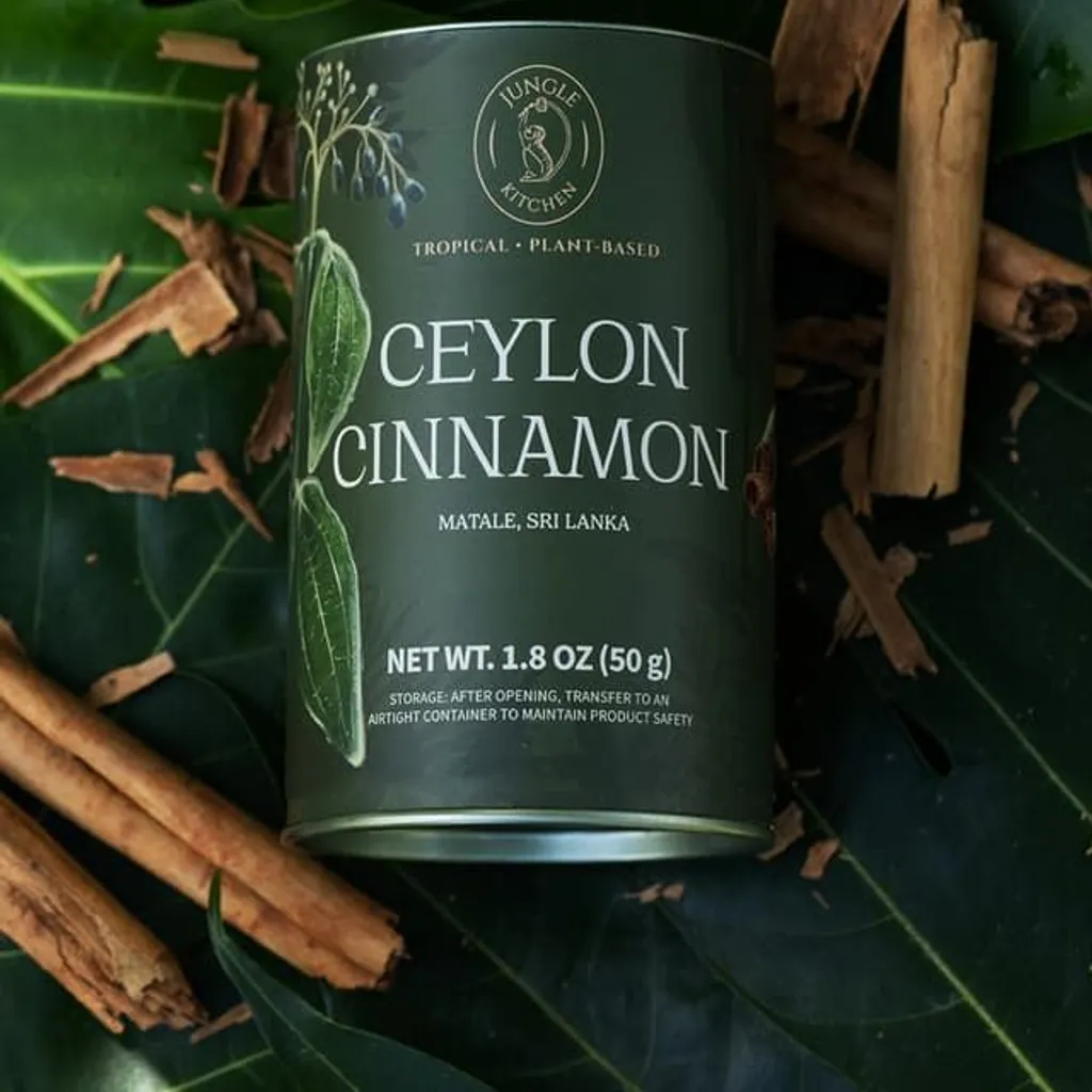 The image features a can of "Ceylon Cinnamon" placed centrally on a bed of green leaves, surrounded by several cinnamon sticks. The lighting is soft and even, highlighting the product's details and rich green color. The texture of the leaves and cinnamon sticks is visible, contributing to a natural, editorial feel.