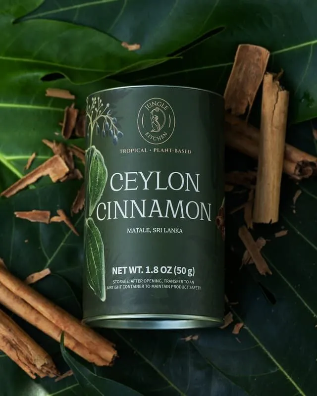 The image features a can of "Ceylon Cinnamon" placed centrally on a bed of green leaves, surrounded by several cinnamon sticks. The lighting is soft and even, highlighting the product's details and rich green color. The texture of the leaves and cinnamon sticks is visible, contributing to a natural, editorial feel.