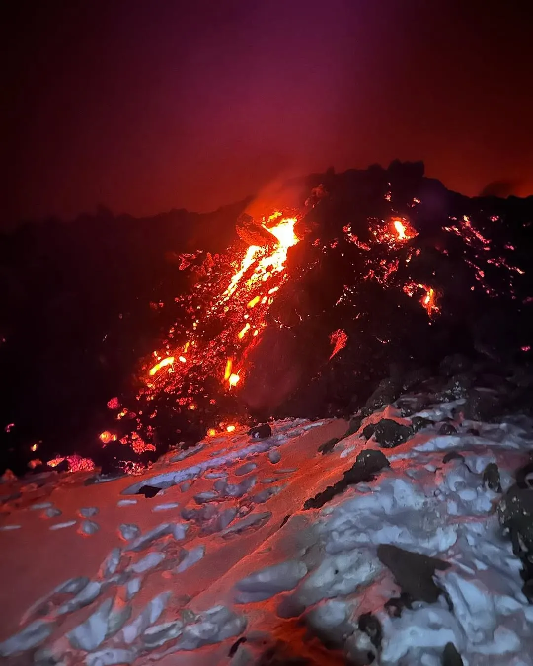 An image captured at night depicting molten lava flowing down a dark, rocky volcanic slope, illuminating the immediate surroundings and casting a reddish glow on patches of snow in the foreground. The sky above is dark with a reddish tinge from the eruption.