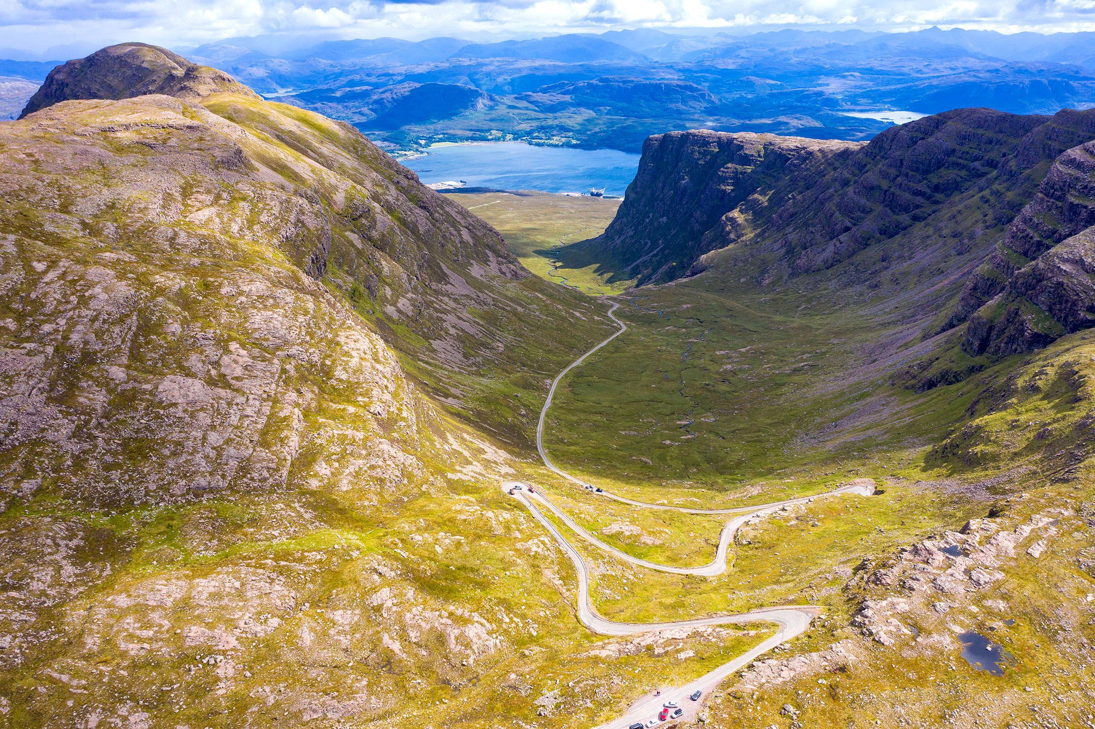 This image is an excellent example of landscape photography, showcasing a dramatic mountain pass with a winding road leading through a valley towards a distant body of water and mountains. The composition is superb, utilizing the road as a strong leading line that draws the eye deep into the scene, creating an impressive sense of depth and scale. The image is exceptionally well-lit with natural light, providing clear visibility and good contrast across the entire landscape without any harsh shadows or blown-out highlights. Sharpness is outstanding, with fine details visible from the foreground rocks to the distant features. Colors are vibrant and appear highly accurate, reflecting the natural hues of the green hills, blue water, and sky. The image is highly photo-realistic, capturing the grandeur of the natural environment. All key elements, including the road, the valley walls, and the distant loch, are in sharp focus, contributing to the overall high quality.