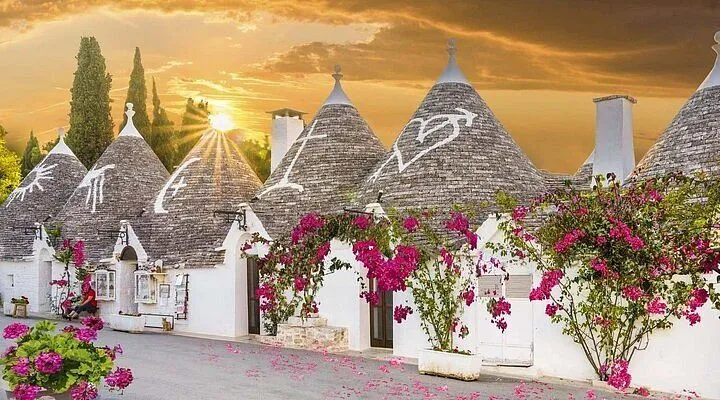 An editorial-style photograph showcasing the iconic Trulli houses of Alberobello, Puglia, at sunset or sunrise. The image features several traditional white buildings with distinctive conical stone roofs, adorned with bright pink bougainvillea. The golden light of the setting or rising sun creates a dramatic and picturesque atmosphere, casting warm tones across the scene.