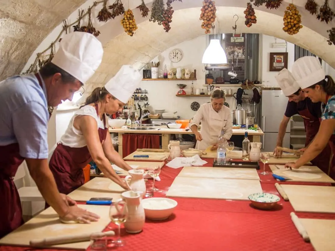 A group of people wearing chef hats and aprons are engaged in a cooking class, rolling dough on a large red table in a rustic kitchen setting.