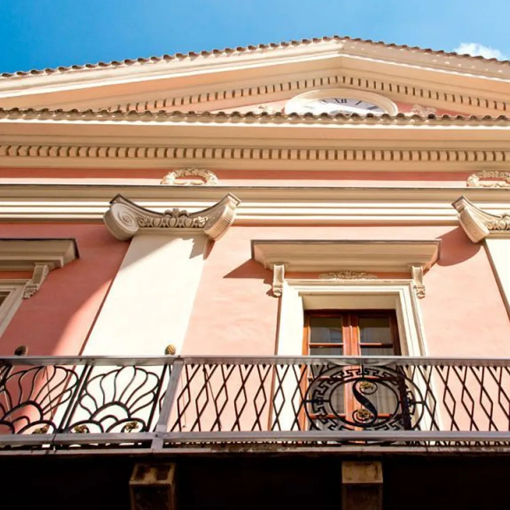 An editorial style image showcasing the exterior of a pink building with ornate architectural details and a decorative balcony. The image captures the upper part of the facade under a clear blue sky.