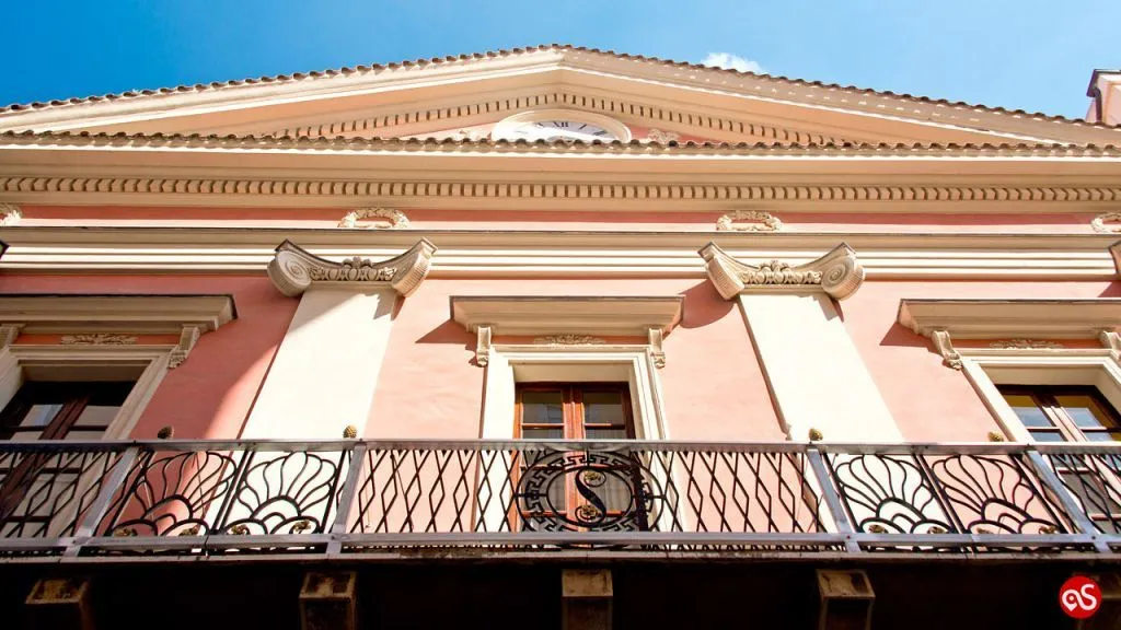 An editorial style image showcasing the exterior of a pink building with ornate architectural details and a decorative balcony. The image captures the upper part of the facade under a clear blue sky.