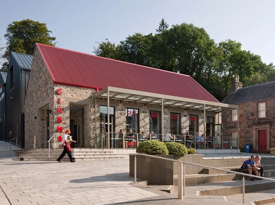 An exterior view of a modern stone building with a prominent red corrugated roof, functioning as a cinema, cafe, and bar, featuring an outdoor seating area with people. The image captures the building clearly under good lighting.