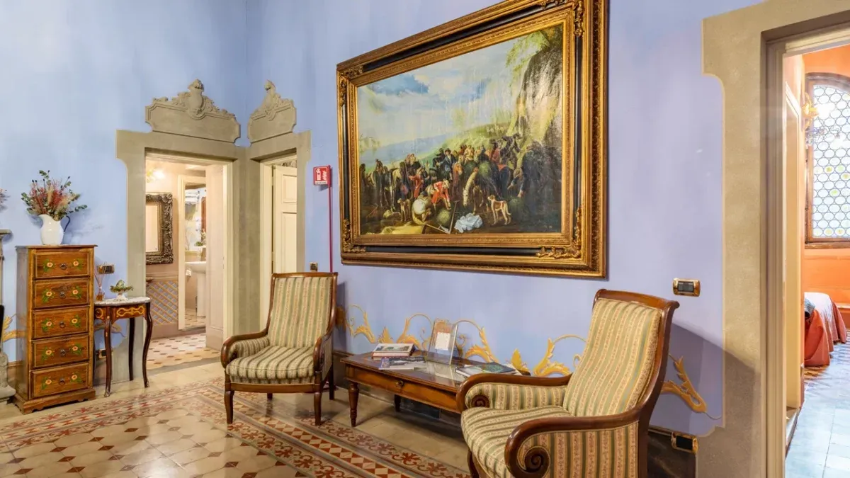 An editorial style image showcasing an elegantly decorated interior space, featuring a large painting on a blue wall, flanked by two classic upholstered chairs. The scene includes a decorative console table and a patterned tiled floor, leading to an open doorway on the left.