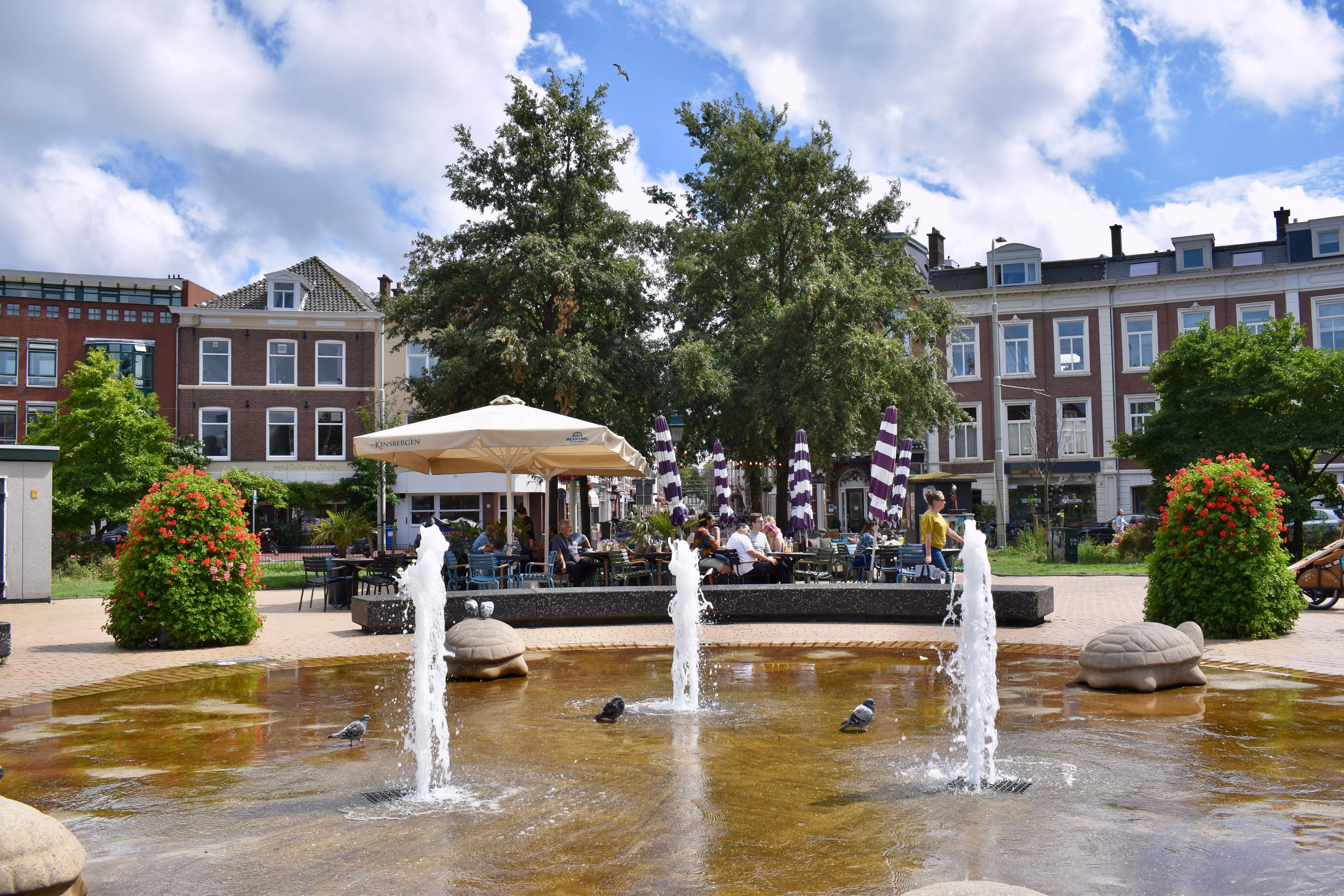 An inviting editorial-style image capturing a lively public square with a fountain in the foreground and an outdoor cafe bustling with people in the midground. Historic buildings and lush trees frame the scene under a bright, partly cloudy sky.