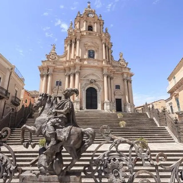 A grand Baroque church, the Chiesa di San Giorgio, stands prominently at the top of a wide staircase, under a bright blue sky with scattered clouds. In the foreground, a bronze equestrian statue is visible, partially framed by an ornate metal railing.