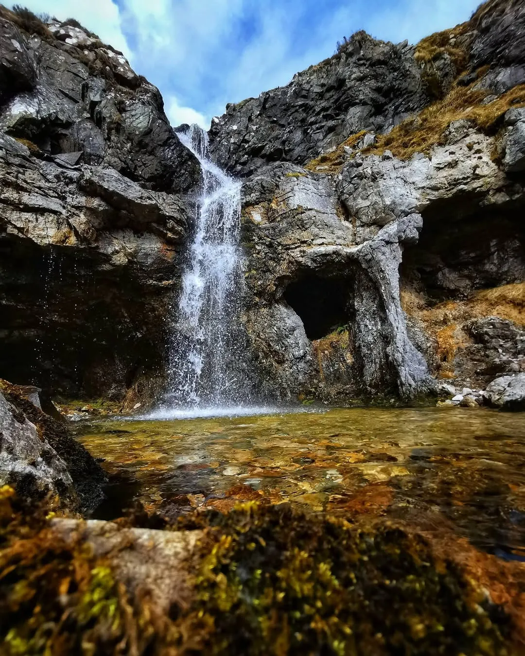 An image depicting a waterfall cascading down a rocky mountain face into a clear pool of water, surrounded by natural rock formations and mossy foreground elements.