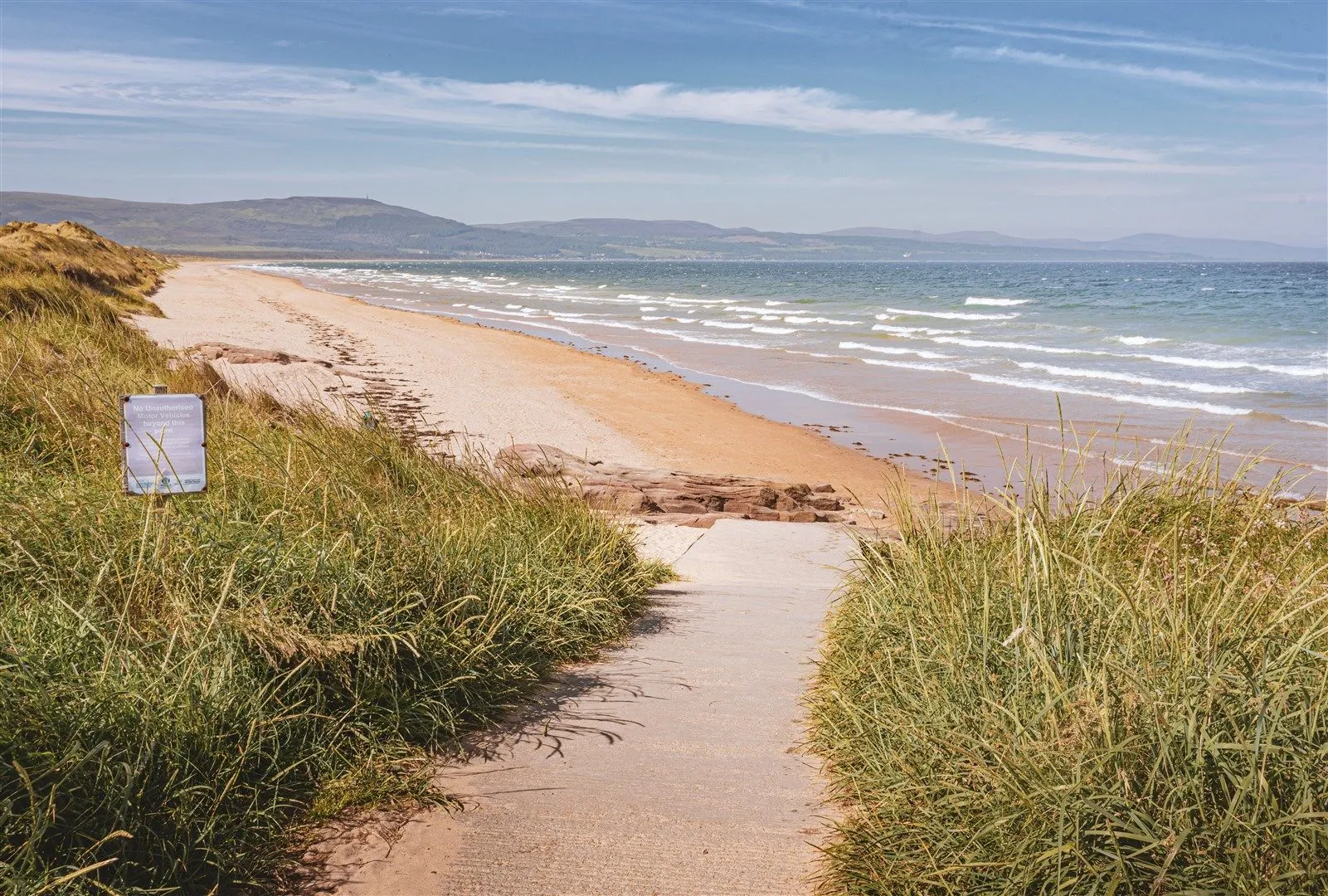 An outdoor shot of a sandy beach under a blue sky with scattered clouds. A concrete path, framed by tall green grass, leads down to the vast expanse of sand and the ocean with gentle waves. Distant mountains are visible on the horizon.