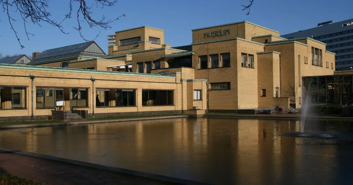 The image captures a modern architectural building, likely a museum, next to a still body of water, possibly frozen or very calm, reflecting the light. The building features light brown brickwork and numerous windows, with a clear blue sky overhead. Bare tree branches are visible in the upper left corner.