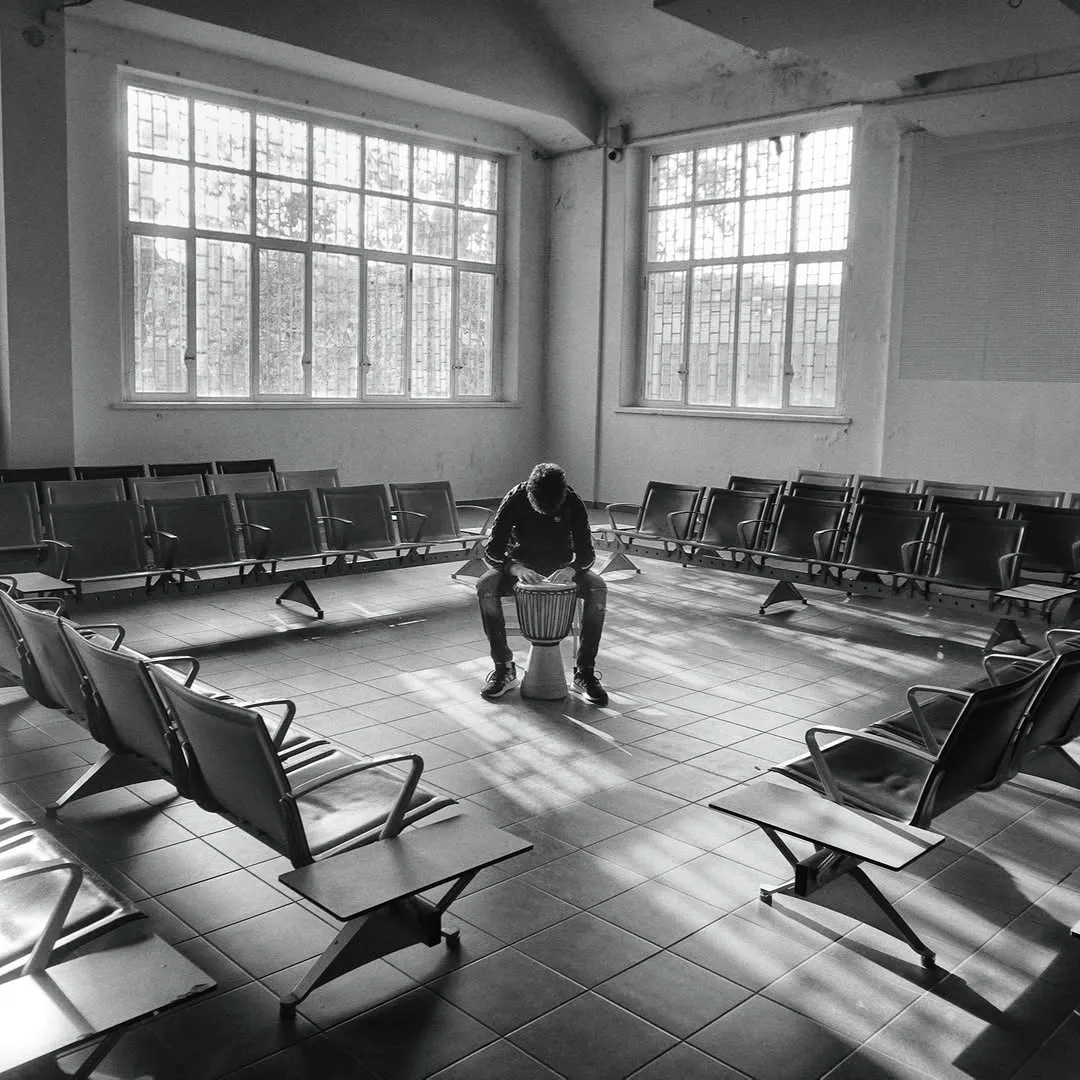 A black and white image depicting a person sitting centrally in a large, empty room, appearing to play a drum. The room is lined with rows of empty chairs arranged in a circular formation, and large windows illuminate the scene with strong natural light, casting dramatic shadows on the tiled floor.