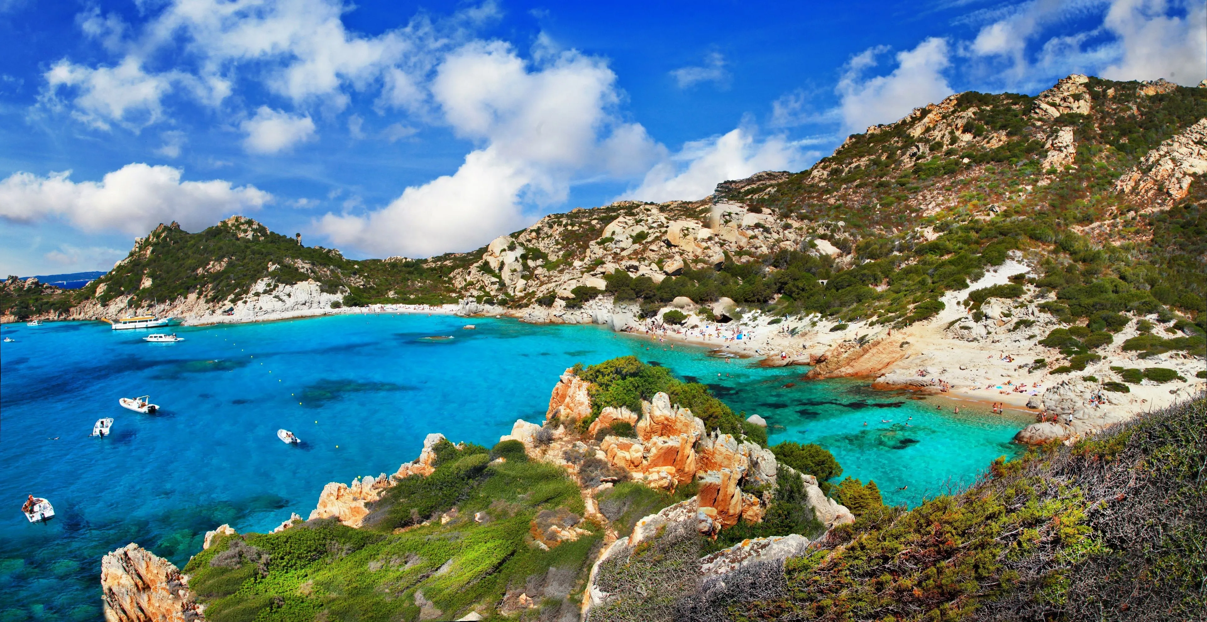 A high-quality image showcasing a beautiful, vibrant blue bay surrounded by rocky, green hills under a clear, bright sky with white clouds. The water is crystal clear, revealing the seabed in shallower areas. A small sandy beach with a few people is visible in the distance.