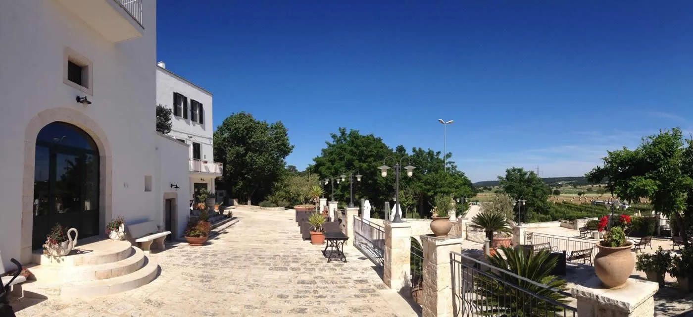 An outdoor scene featuring a white building with balconies on the left, a stone-paved pathway in the foreground, and lush green trees and streetlights leading towards the background under a clear blue sky. Various potted plants and ornate fences are also visible.