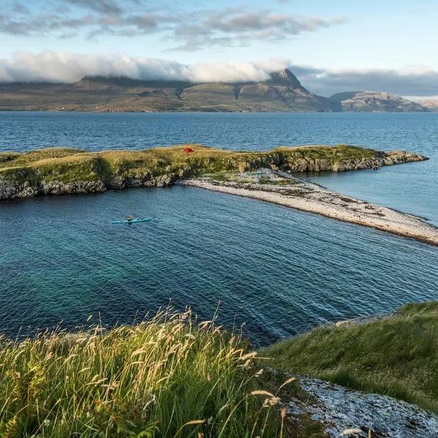 A scenic view of a small, grassy island connected by a natural gravel causeway to another landmass, surrounded by clear blue-green water. A person in a blue kayak paddles in the calm water. Distant mountains, partially covered by clouds, are visible under a bright, partly cloudy sky.