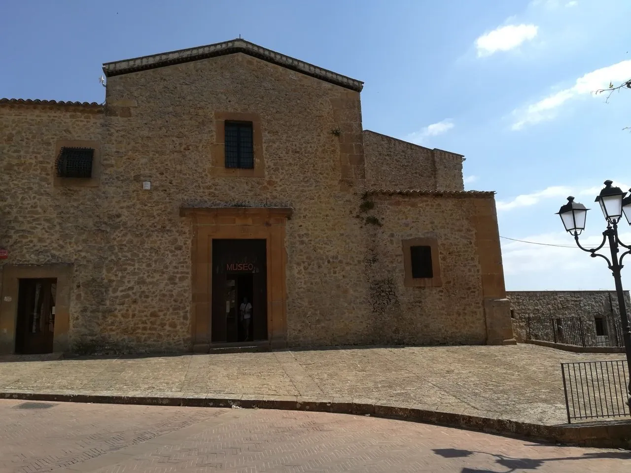 An editorial image of a historic stone building, identified as a 'MUSEO' (museum) by the sign above its main entrance. The building is constructed of light-colored stone blocks, featuring a central entranceway and windows. The foreground shows a paved plaza or courtyard area leading up to the building. The sky above is clear and blue with some scattered clouds, indicating a sunny day.