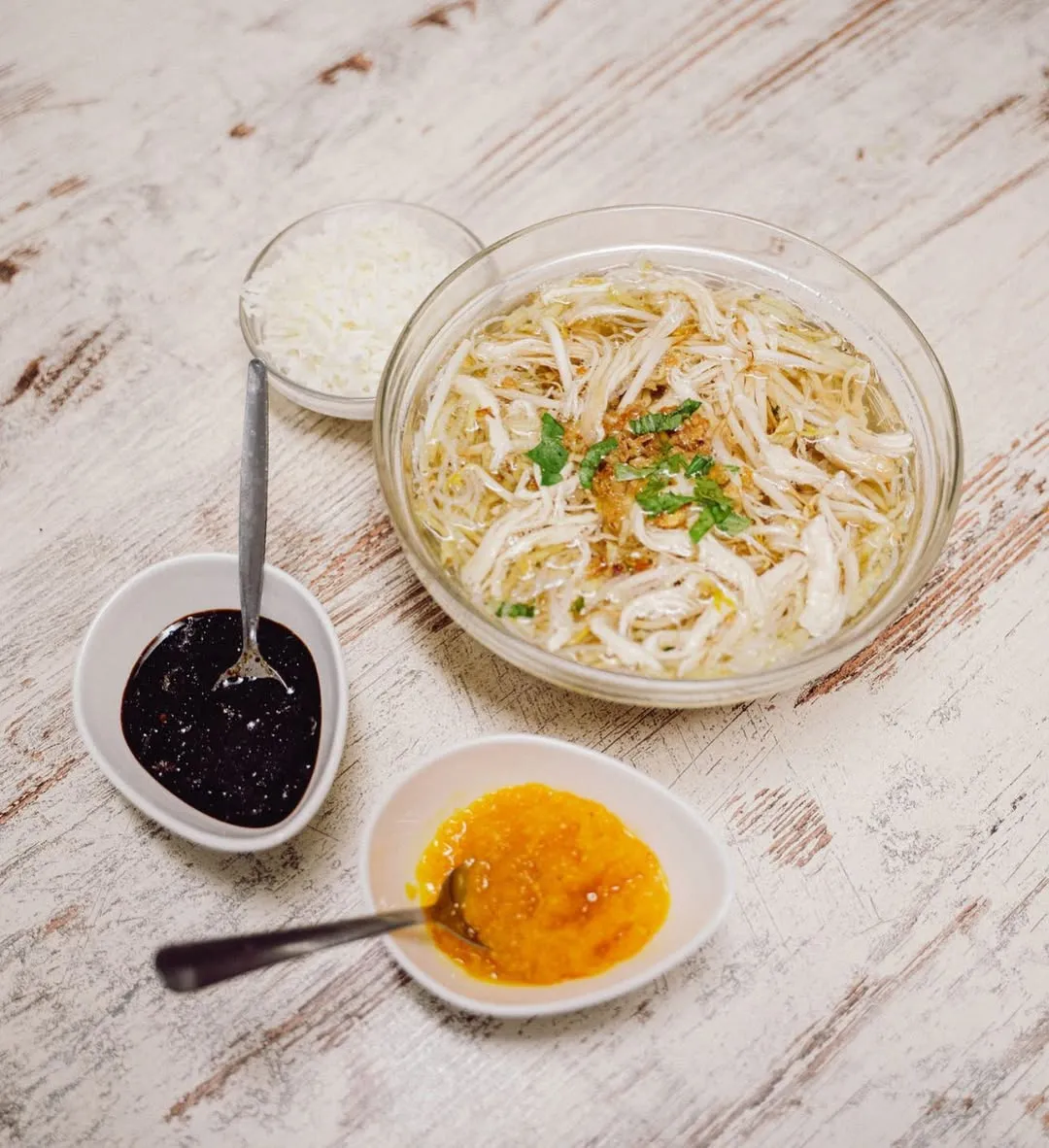 A well-composed and well-lit image featuring a clear bowl of chicken noodle soup, possibly Soto Ayam, garnished with shredded chicken, fried shallots, and herbs. It is accompanied by two smaller white bowls containing dark sauce and a yellow chili paste, all set on a light wooden table.
