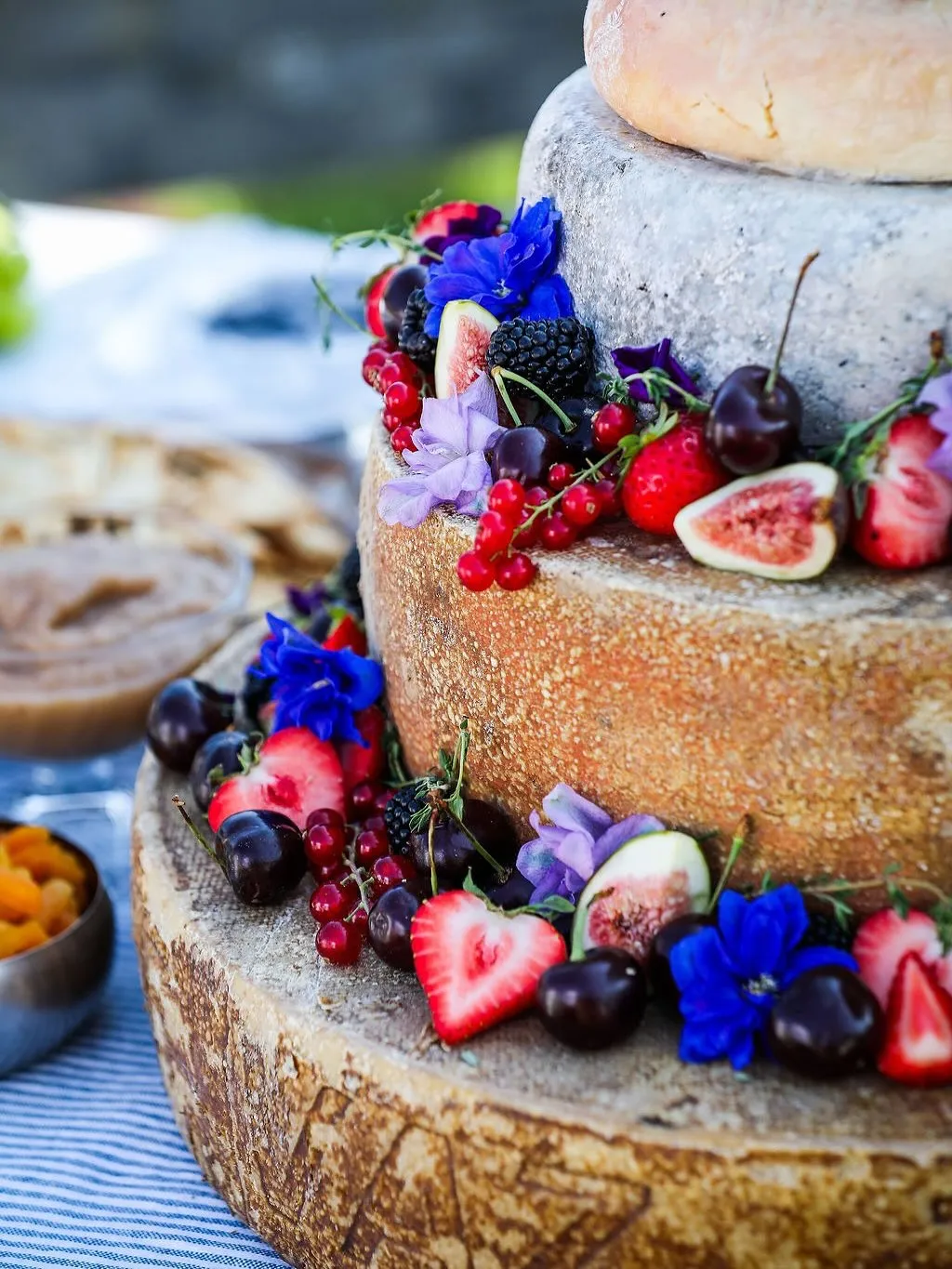 A vibrant, close-up image of a tiered cheese wheel cake elegantly adorned with an assortment of fresh berries, figs, cherries, and colorful edible flowers, set outdoors with a softly blurred background.