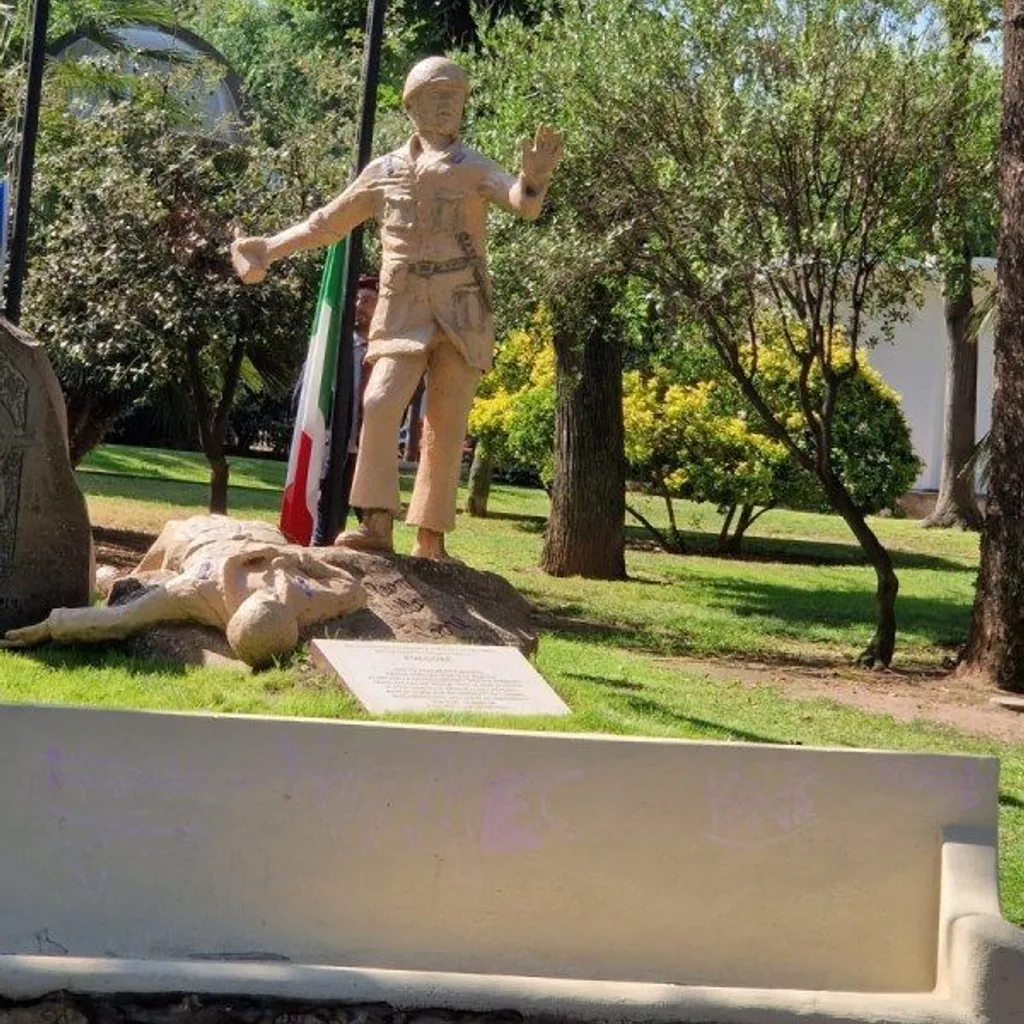A photograph of a war memorial or monument featuring two statues in a park-like setting. One statue depicts a standing soldier, while another figure lies on the ground in front of him. Green trees and grass surround the statues, with a partially visible Italian flag in the background.