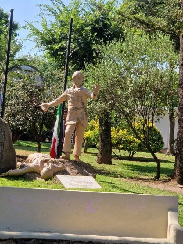 A photograph of a war memorial or monument featuring two statues in a park-like setting. One statue depicts a standing soldier, while another figure lies on the ground in front of him. Green trees and grass surround the statues, with a partially visible Italian flag in the background.
