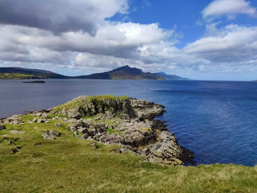 A picturesque view of a rocky coastline extending into a vast blue sea, with distant mountains under a partly cloudy sky.