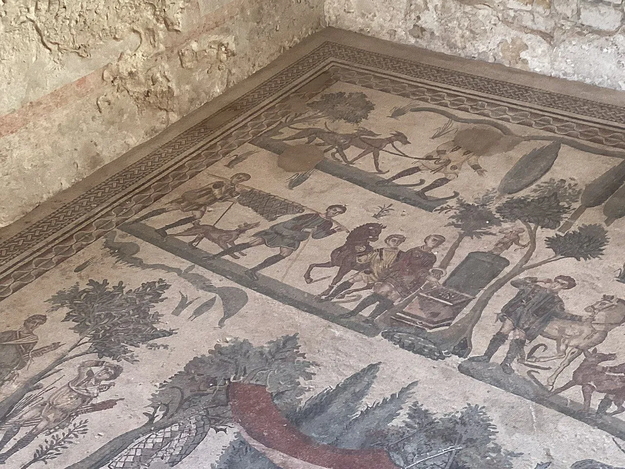 An eye-level shot of an ancient Roman mosaic floor, depicting a hunting scene with human figures, horses, and dogs in a natural setting. The mosaic is intricate, showcasing various earthy tones and faded colors due to age. A rough, ancient-looking stone wall is visible in the upper left corner, indicating the setting is within ruins or a historical site.