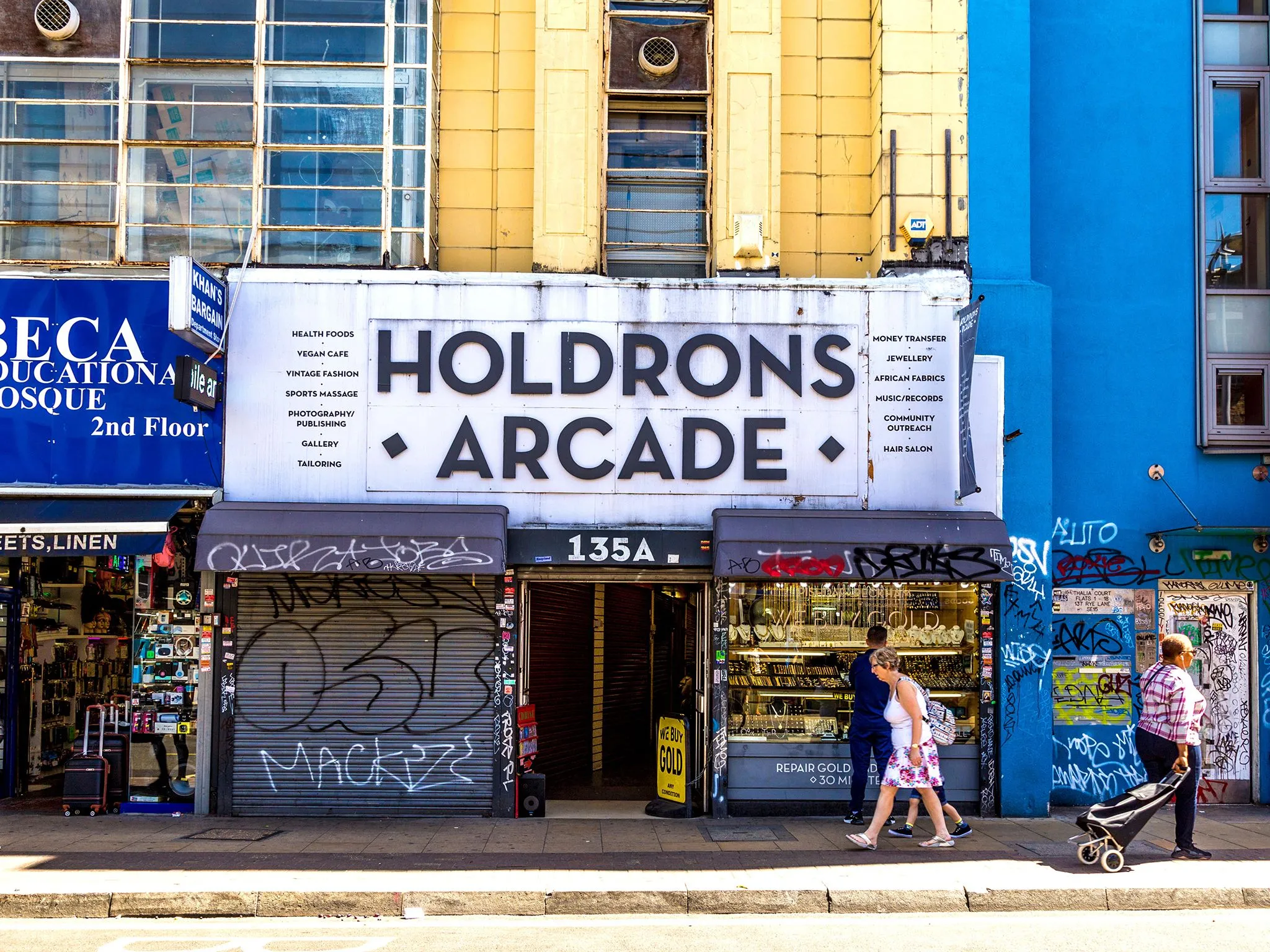 The image showcases an urban street scene featuring the facade of "HOLDRONS ARCADE." It is well-lit with natural light, providing good color accuracy and a strong sense of photo-realism. Key elements like the main signage and shop fronts are sharp and in focus. The composition is generally balanced, though the presence of graffiti and numerous small signs contributes to a somewhat busy aesthetic.