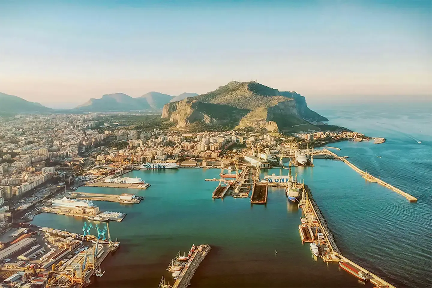 An aerial view of a bustling port city nestled at the base of a large mountain, with numerous ships, docks, and urban sprawl extending towards the distant horizon.