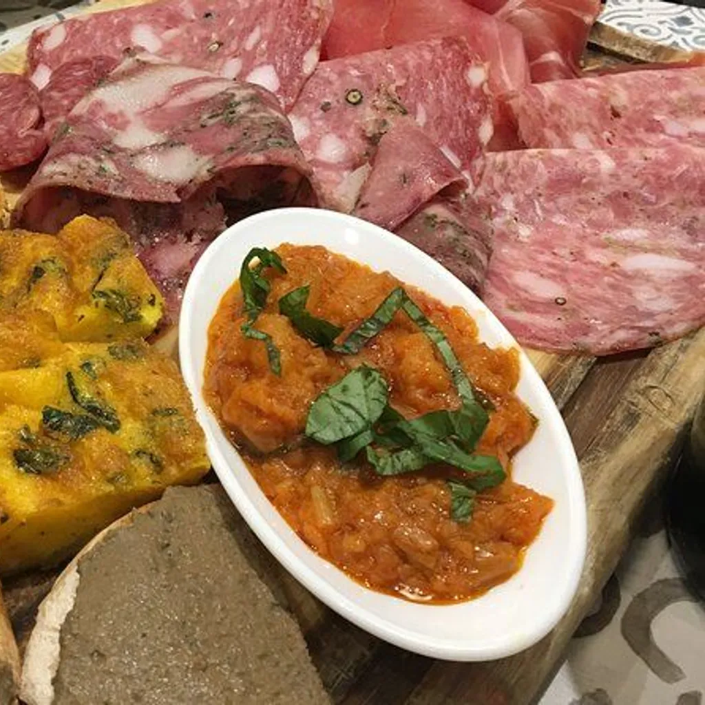 A close-up shot of an appetizer platter featuring various items such as sliced charcuterie, a yellow frittata-like dish, a bowl of red sauce, and bread with spread. The composition fills the frame, presenting a rich culinary display.