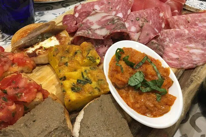 A close-up shot of an appetizer platter featuring various items such as sliced charcuterie, a yellow frittata-like dish, a bowl of red sauce, and bread with spread. The composition fills the frame, presenting a rich culinary display.