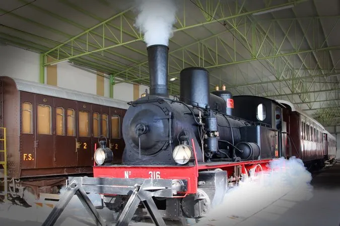 The image features a vintage steam locomotive indoors, emitting smoke. It is well-composed, with the train centrally framed and capturing significant details. The focus is sharp on the main subject, contributing to a high degree of photo-realism. Colors appear accurate, reflecting the dark tones of the locomotive and the industrial setting.