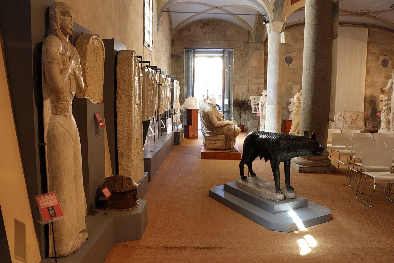 An editorial style image capturing the interior of a museum hall, showcasing ancient sculptures and stone reliefs. A prominent bronze statue of a wolf stands in the foreground, with various artifacts displayed along the walls. Natural light illuminates the scene, highlighting the architectural features and exhibits.