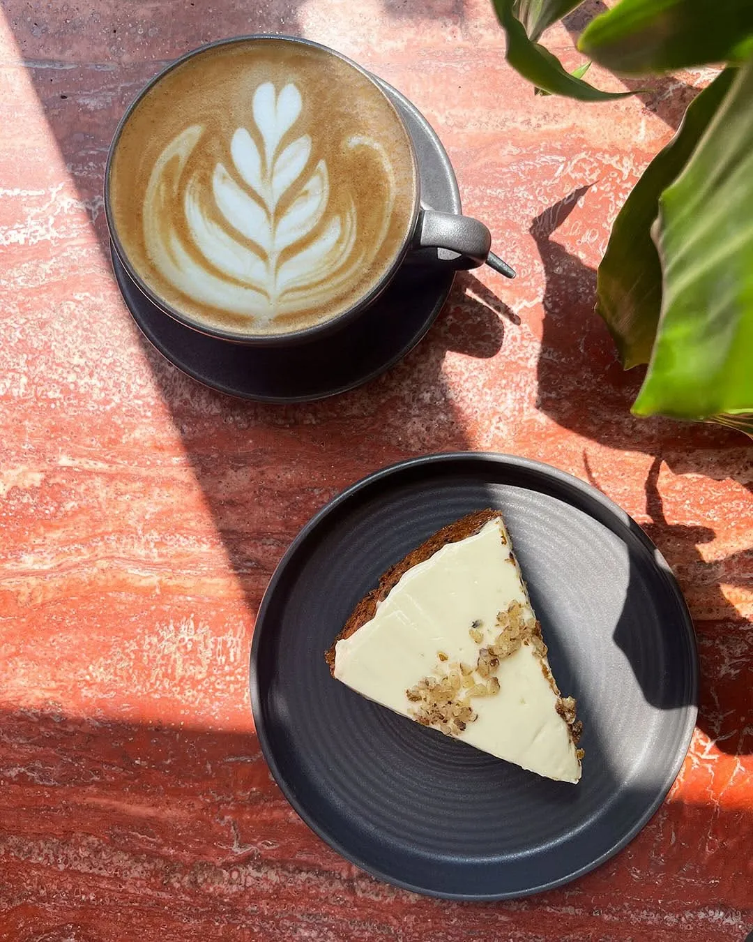 An overhead shot of a latte with detailed latte art and a slice of cake on a dark plate, set on a reddish marble-like table. Strong natural light casts prominent shadows across the scene.