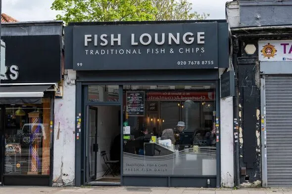 An exterior shot of 'Fish Lounge' a traditional fish & chips shop. The storefront features a dark grey facade with white lettering for the shop name and services. The entrance is open, revealing a glimpse of the interior with some customers and staff. The street pavement is visible in the foreground, and some tree foliage is at the top.