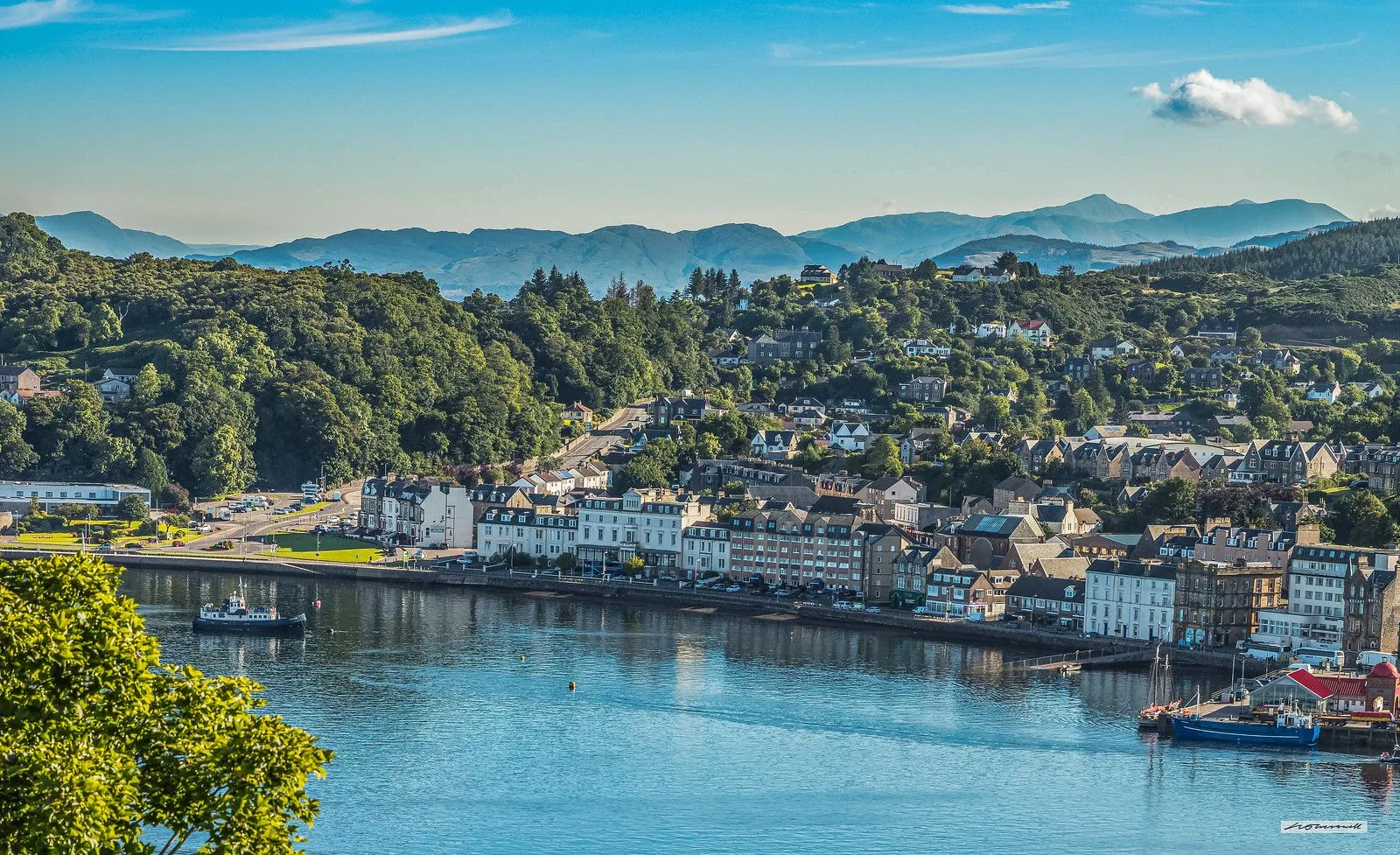 An editorial quality image depicting a scenic town nestled along a body of water, backed by lush green hills and distant mountains under a clear blue sky.