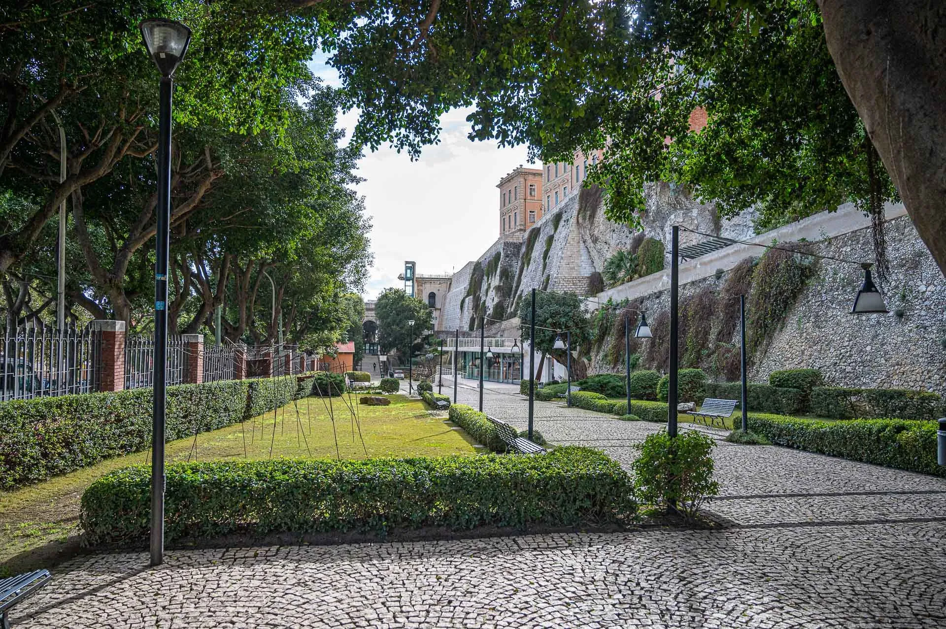 An inviting and serene view of an urban park with a cobbled pathway leading into the distance, framed by lush green trees. A large stone wall with a historic building atop it forms the background.