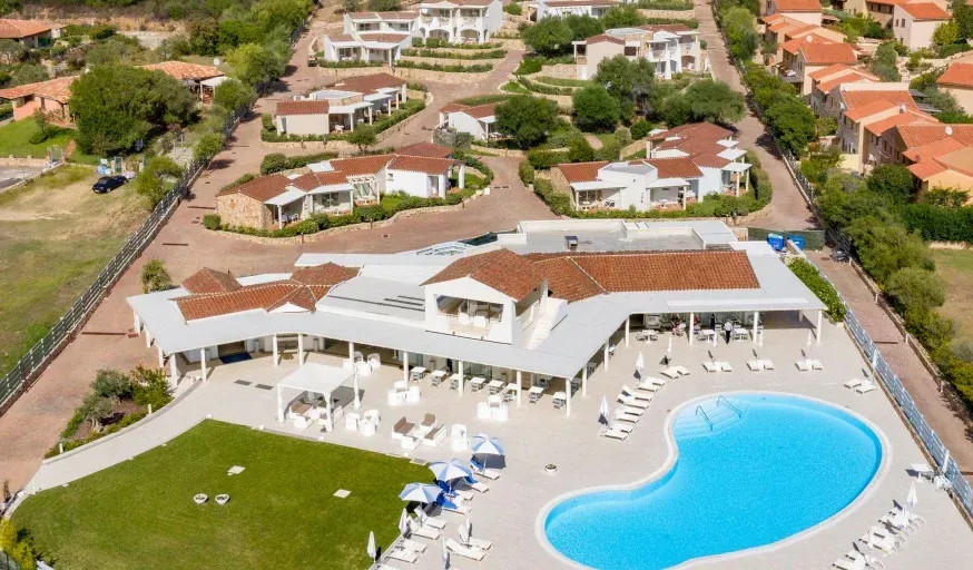 An aerial shot of a resort complex featuring a large main building with terracotta roofs, an outdoor dining area, a large swimming pool, and numerous white villas scattered on terraced hillsides. The landscape includes green lawns, trees, and paved pathways.