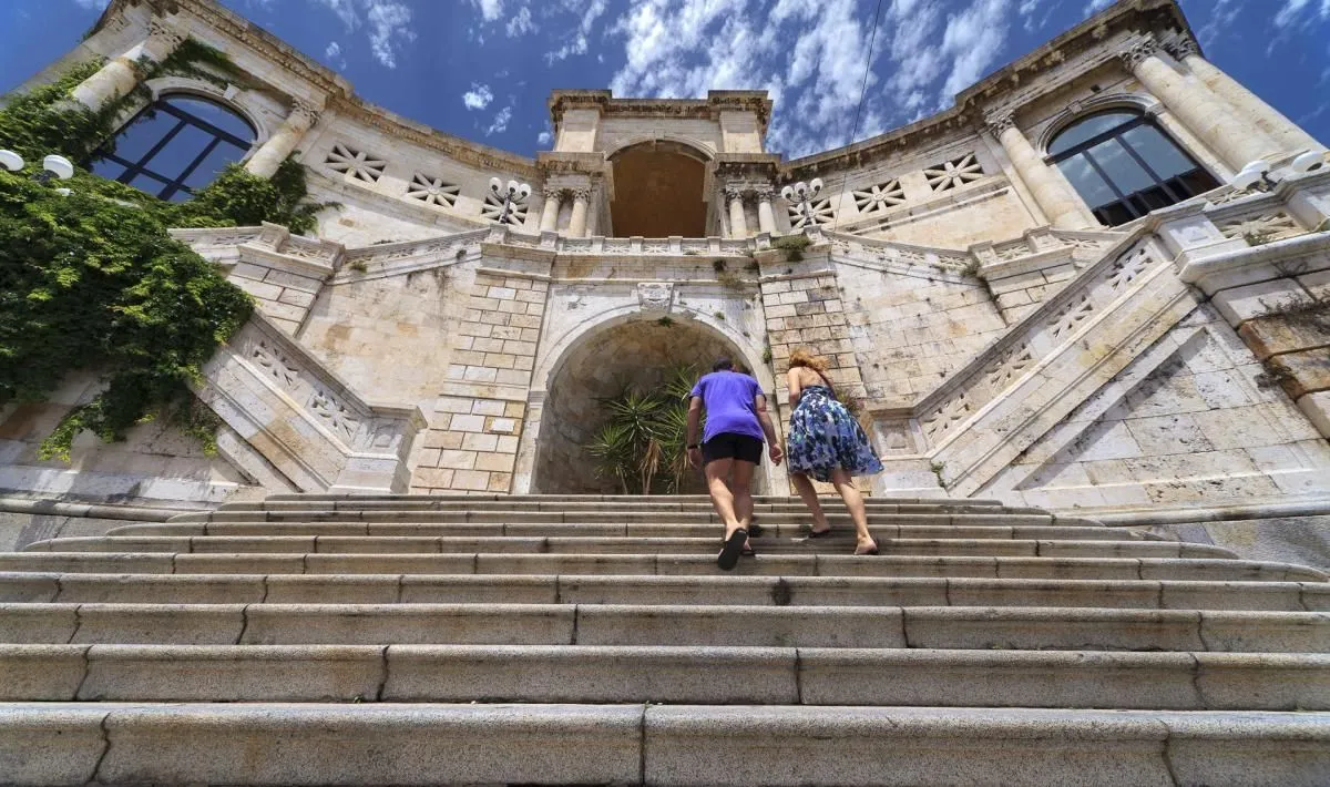 A low-angle shot captures two people ascending a grand, ornate stone staircase leading up to an impressive historical building under a bright blue sky with scattered clouds. The architectural details of the structure are prominent.