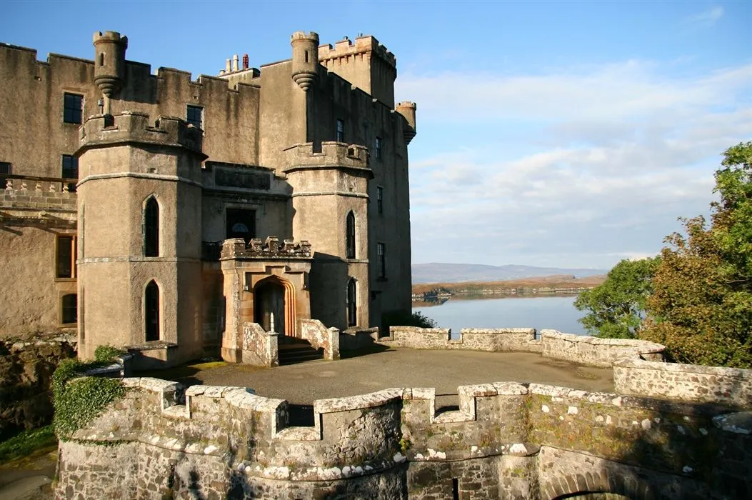 An image of a historic stone castle situated next to a large body of water under a clear blue sky with some clouds.