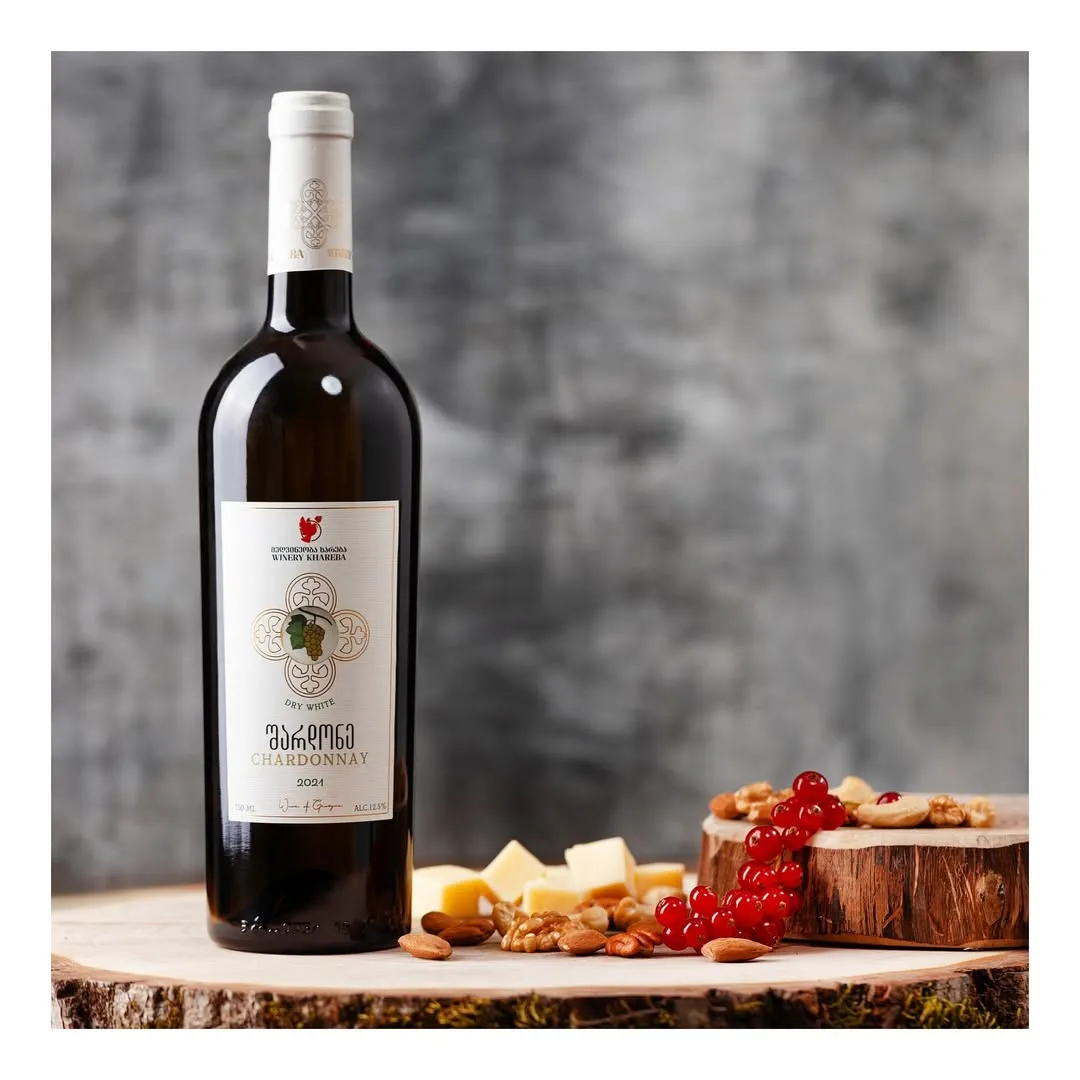 A professionally captured still life image featuring a bottle of white wine, accompanied by cheese cubes, almonds, walnuts, and redcurrants, all artfully arranged on wooden surfaces. The background is a blurred, dark grey texture.