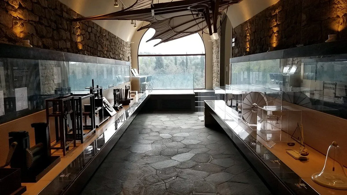 An interior view of a museum or exhibition space, featuring a stone floor, display cases with various items, and a large arched window offering a view of greenery outside. An interesting, possibly artistic, structure hangs from the ceiling.