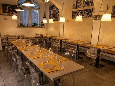 An interior shot of a restaurant dining area, showcasing a long table set with glasses and placemats, along with other smaller tables in the background. The space is illuminated by hanging lamps and natural light from a window.