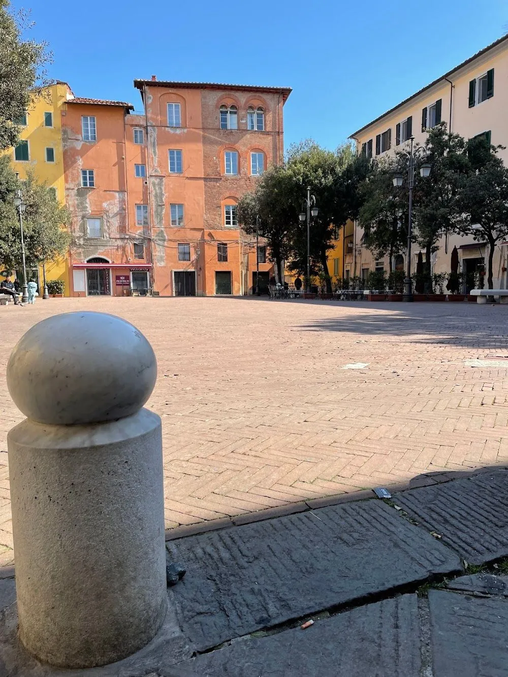 An editorial-style image capturing a sunny piazza surrounded by historic buildings. The foreground features a marble bollard, leading the eye into the expansive brick-paved square. The buildings are primarily terracotta and yellow, under a clear blue sky, with scattered trees providing shade. The scene depicts a quiet urban setting.