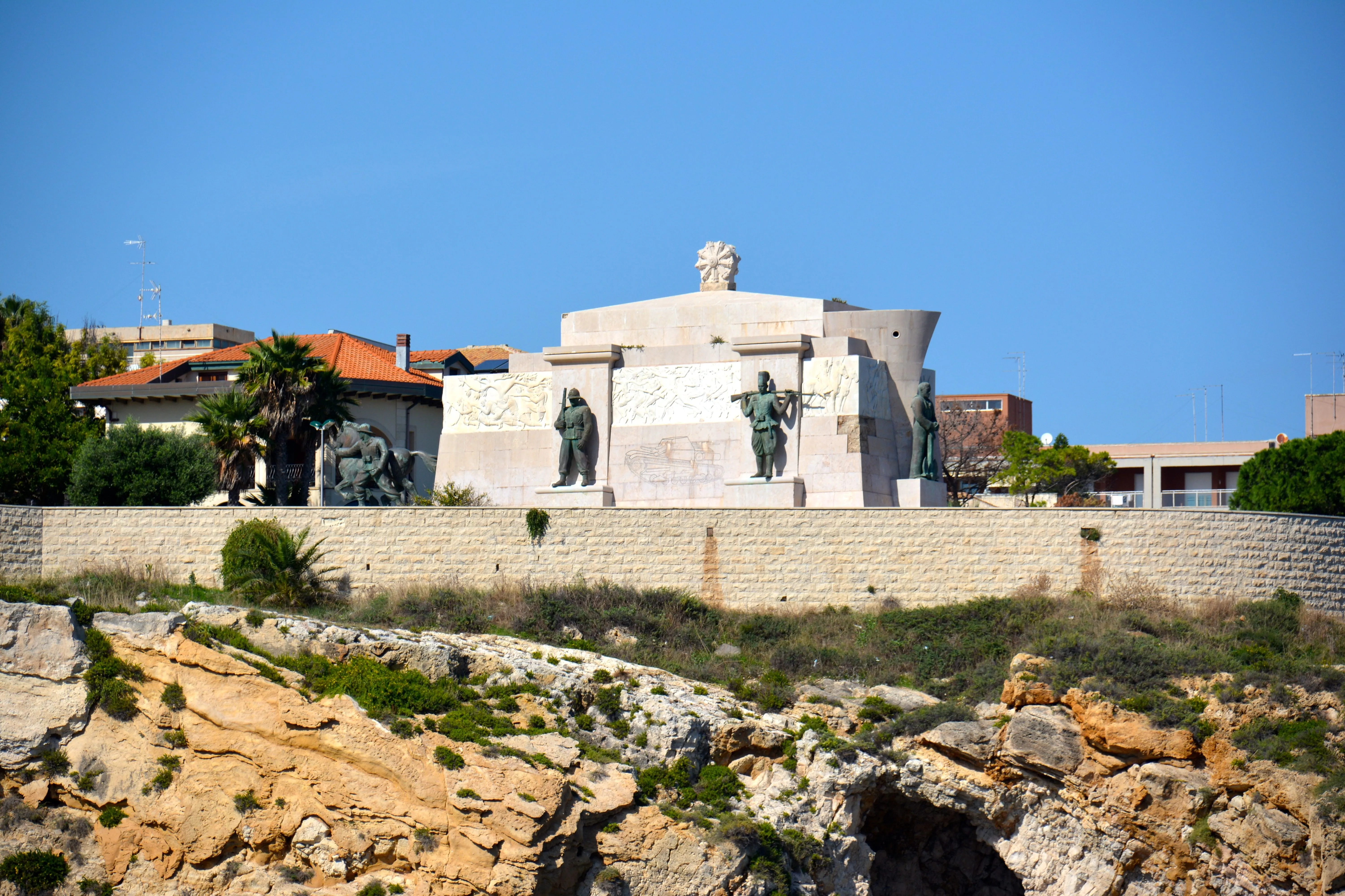An outdoor daytime shot of a large stone monument featuring multiple statues, set on a raised platform above a rocky, vegetated cliffside under a clear blue sky. The monument appears to be a historical or commemorative structure.