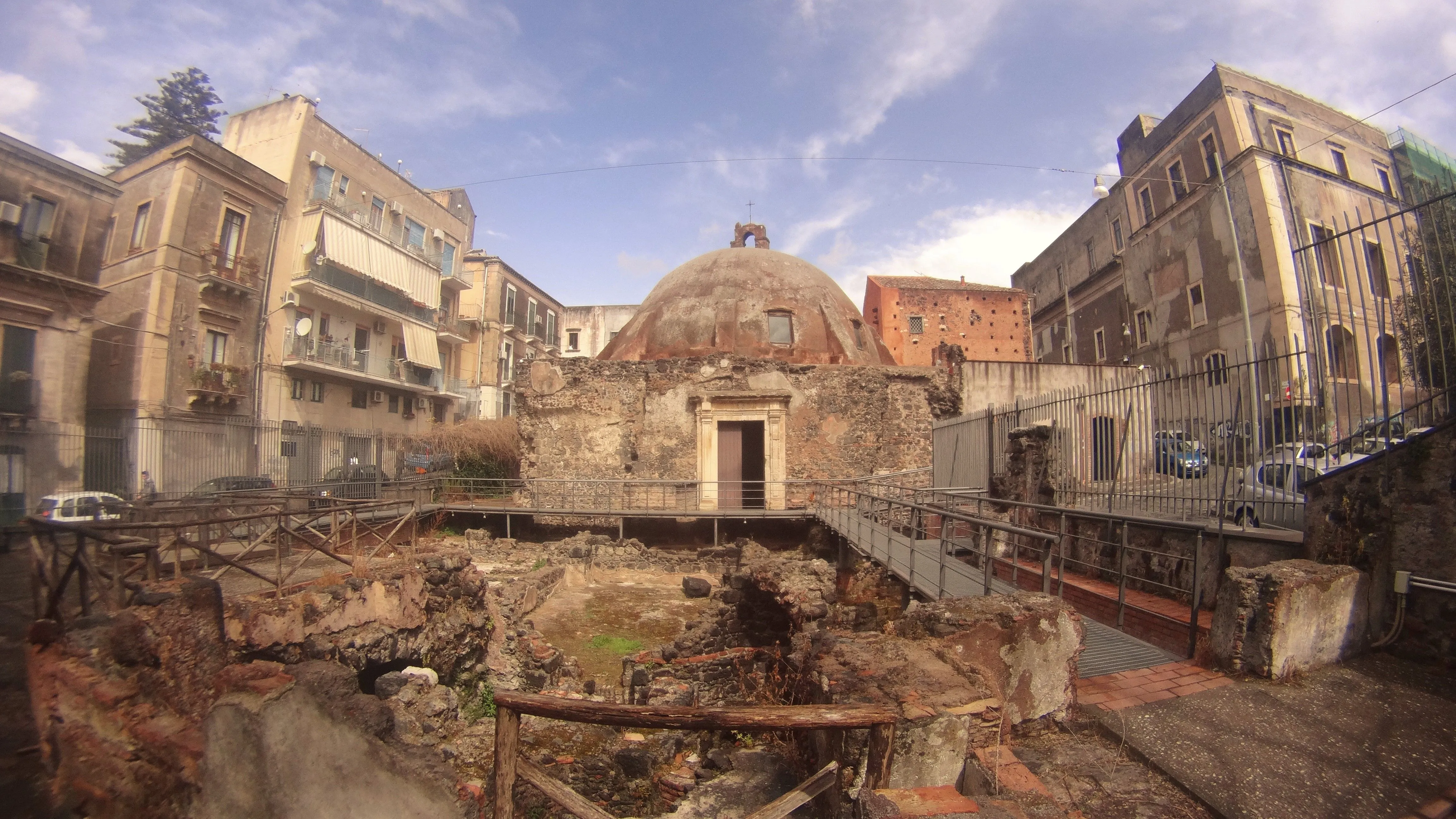 The image captures an ancient domed structure amidst archaeological ruins, framed by surrounding city buildings and modern walkways.