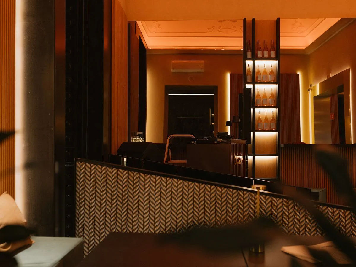 An interior shot of a stylish bar or lounge, characterized by its warm orange lighting and modern design elements. The image features a distinctive patterned wall and illuminated shelving displaying bottles, creating an atmospheric and inviting ambiance. The composition is decent, capturing various elements of the space, although some areas are quite dim.