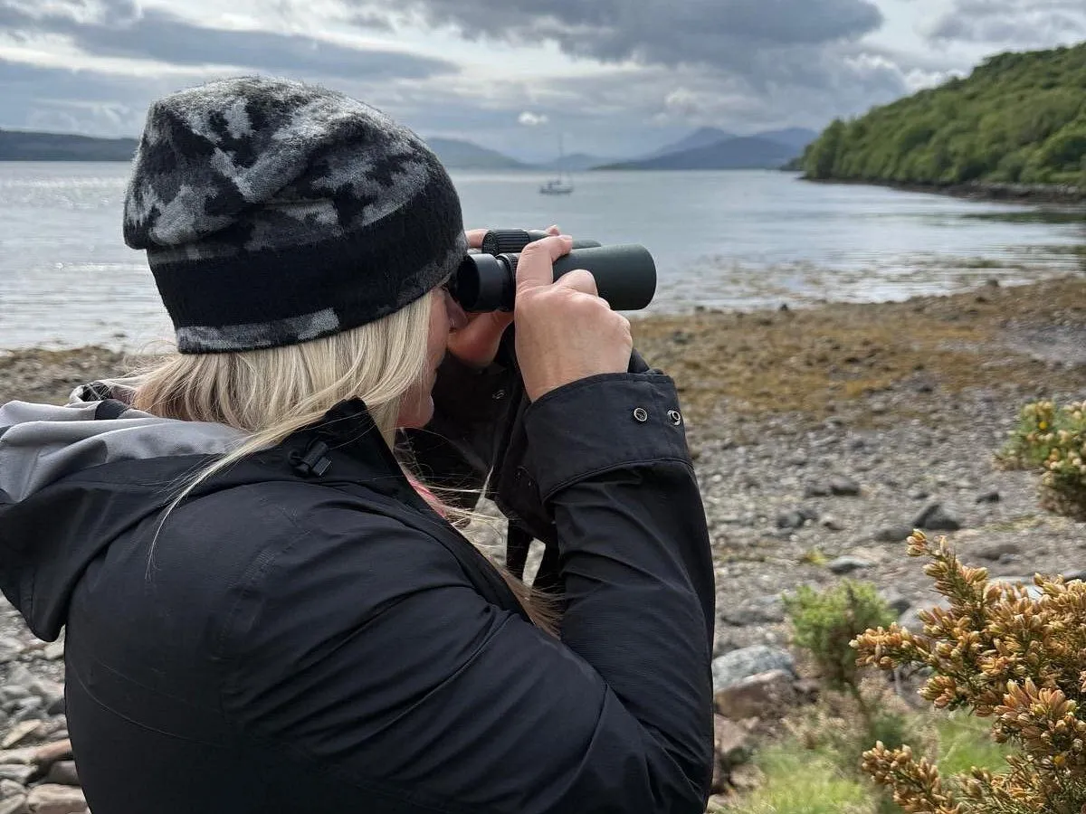 An image captured from behind of a person on a rocky shoreline, looking out over a body of water with binoculars. Distant mountains and a boat are visible in the background under an overcast sky.