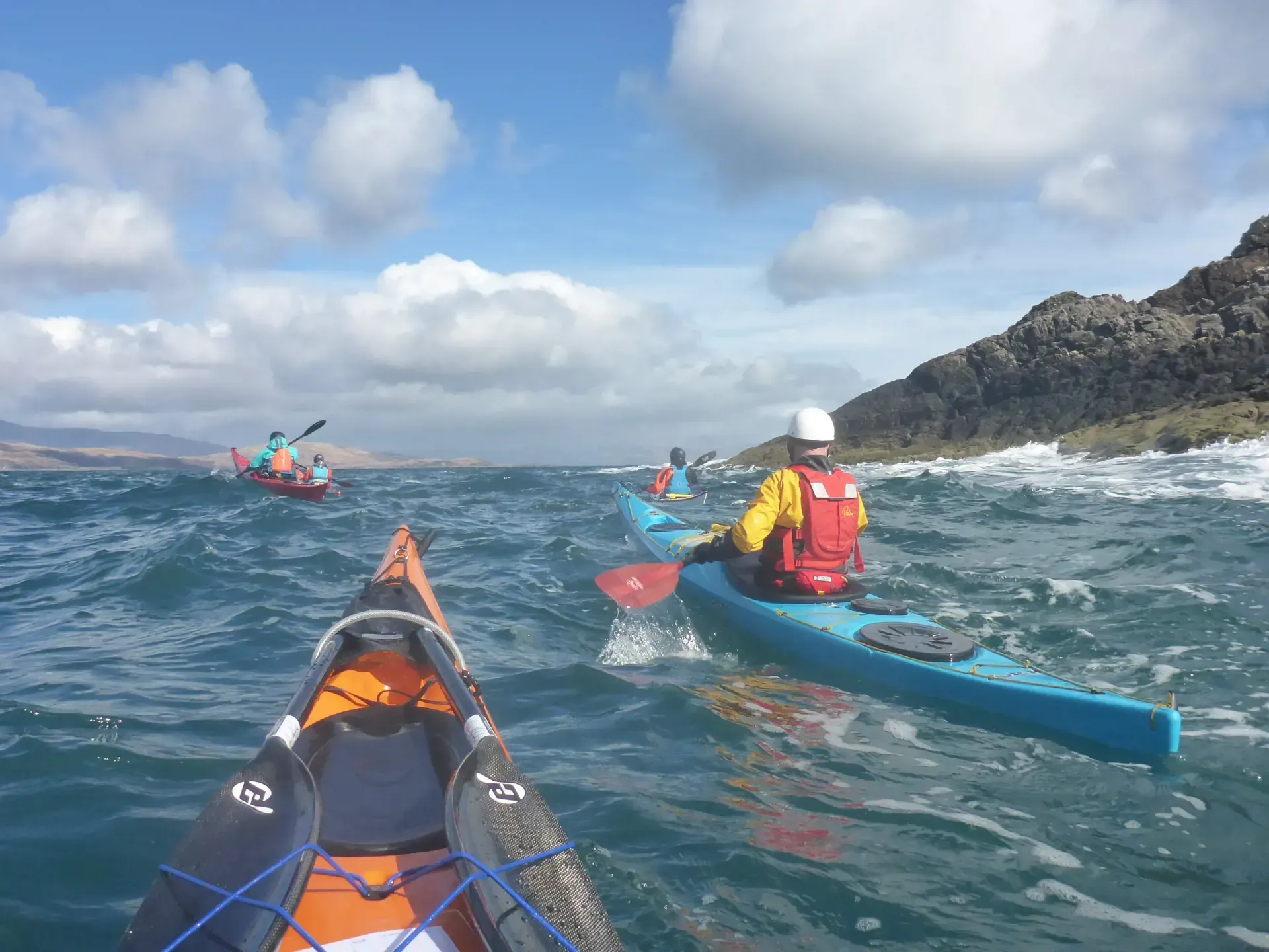 A dynamic outdoor shot featuring several kayakers navigating choppy ocean waters under a partly cloudy sky, with distant land visible.