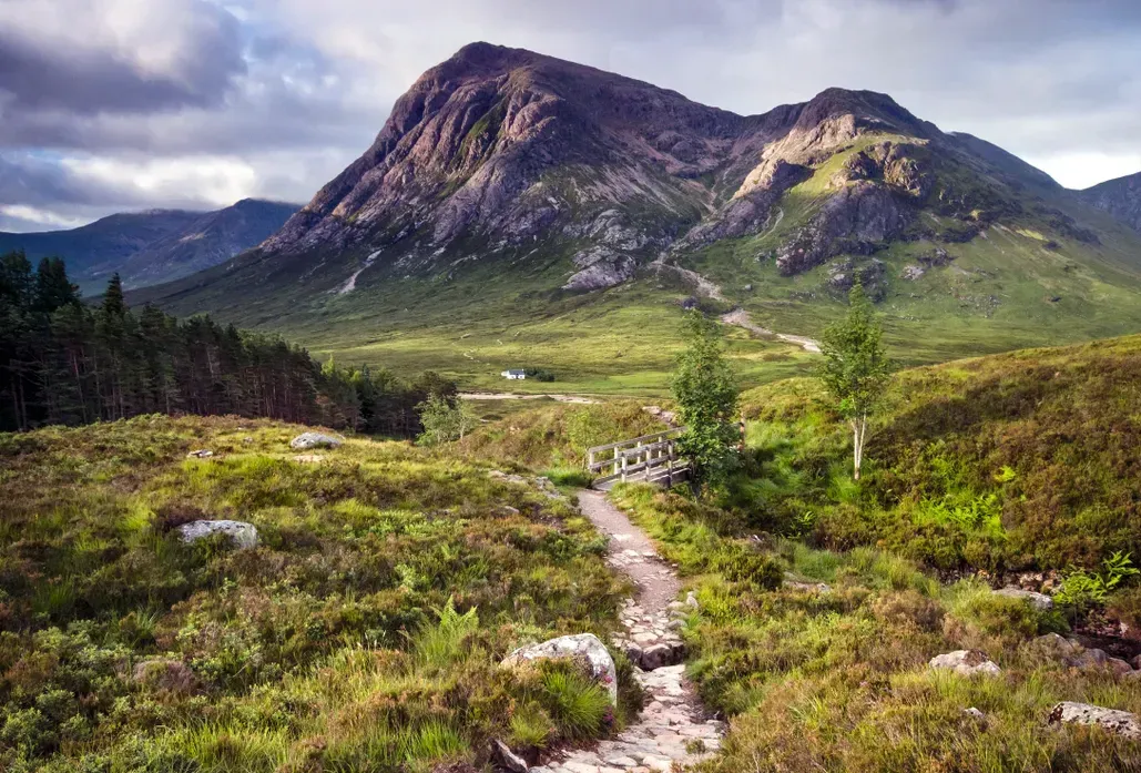 A beautiful landscape photograph depicting a winding stone path leading towards a small wooden bridge and a distant white house, all set against the backdrop of a majestic, rugged mountain. The foreground is rich with green and brown moorland vegetation under a dynamic sky.